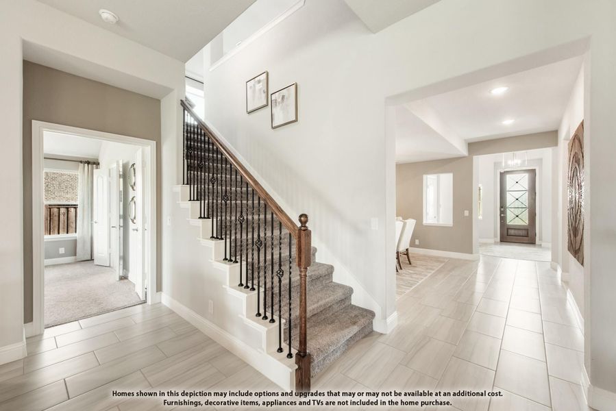 Bright home entryway with carpeted staircase, wood and iron railing, and light tile floors leading to front door.