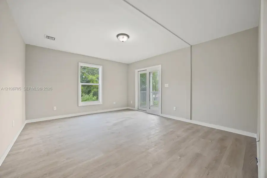 Neutral-toned interior featuring wood-finish flooring, a glass-pane sliding door with exterior access, a single window, and a flush-mount ceiling light fixture Neutral-toned interior featuring wood-finish flooring, a glass-pane sliding door with exterior access, a single window, and a flush-mount ceiling light fixture
