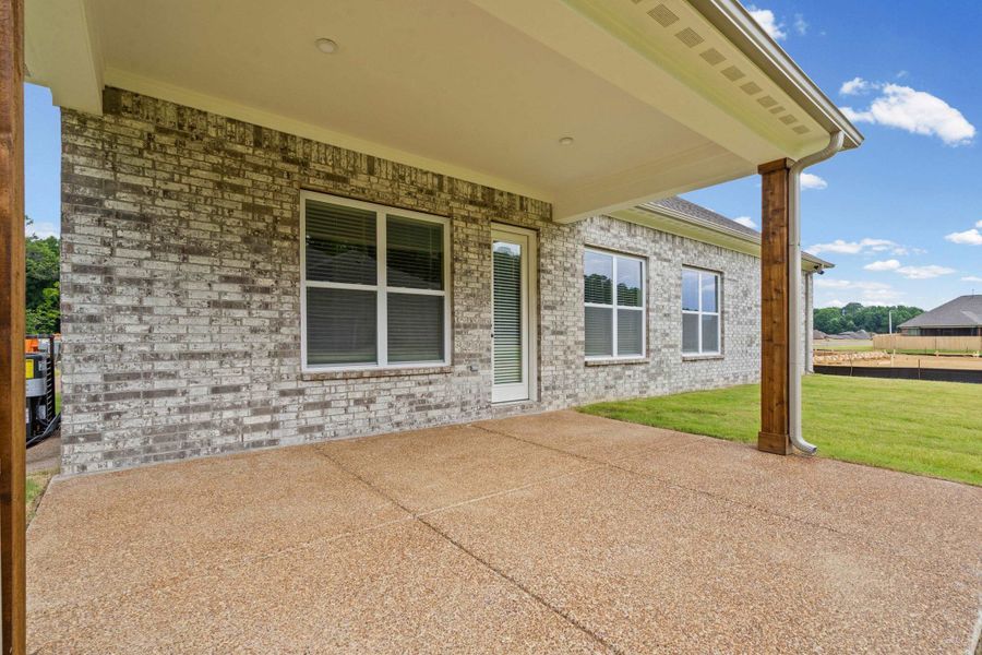 Covered patio featuring a textured concrete surface, brick exterior, white framed windows, and a wood-wrapped support column Covered patio featuring a textured concrete surface, brick exterior, white framed windows, and a wood-wrapped support column