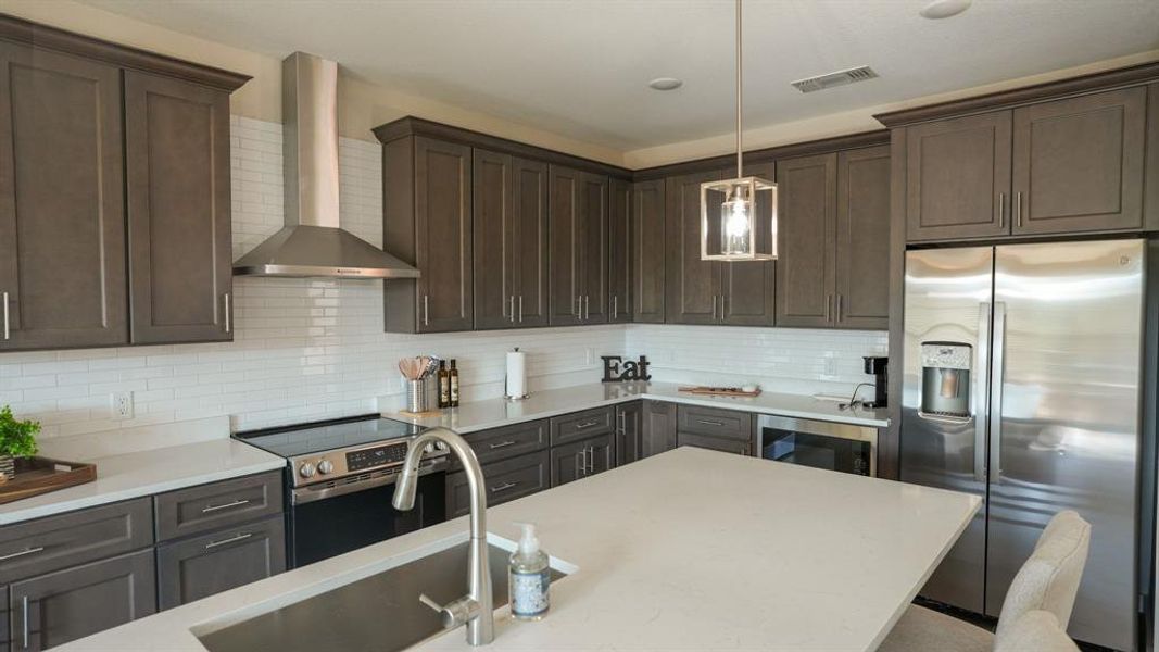 Kitchen featuring appliances with stainless steel finishes, wall chimney range hood, dark brown cabinets, pendant lighting, and light stone counters