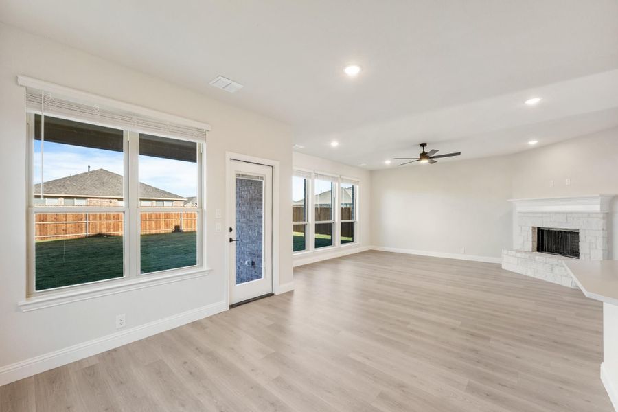 Open living room with light wood floors, stone fireplace, ceiling fan, and backyard-facing windows Open living room with light wood floors, stone fireplace, ceiling fan, and backyard-facing windows