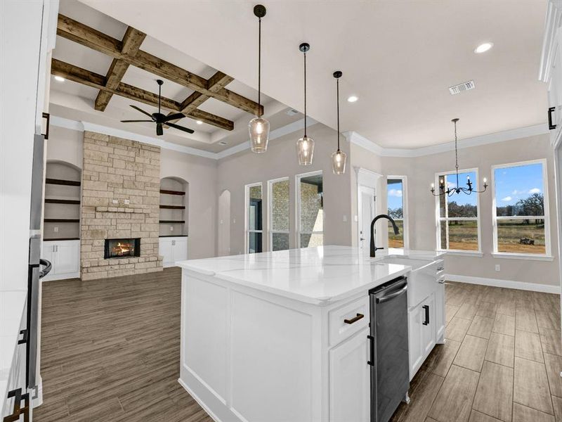 Kitchen featuring coffered ceiling, a stone fireplace, an island with sink, light stone counters, and built in shelves