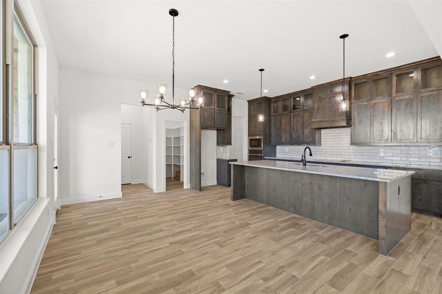 Kitchen featuring backsplash, dark brown cabinetry, light wood-style floors, a chandelier, and a large island with sink Kitchen featuring backsplash, dark brown cabinetry, light wood-style floors, a chandelier, and a large island with sink
