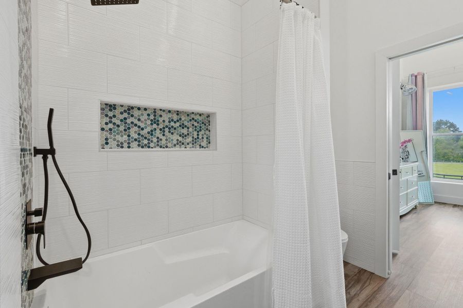 This bathroom features a modern tub with a sleek rainfall showerhead and a hexagonal tile accent. A white shower curtain adds elegance, and the room opens to a bright space with wooden flooring.