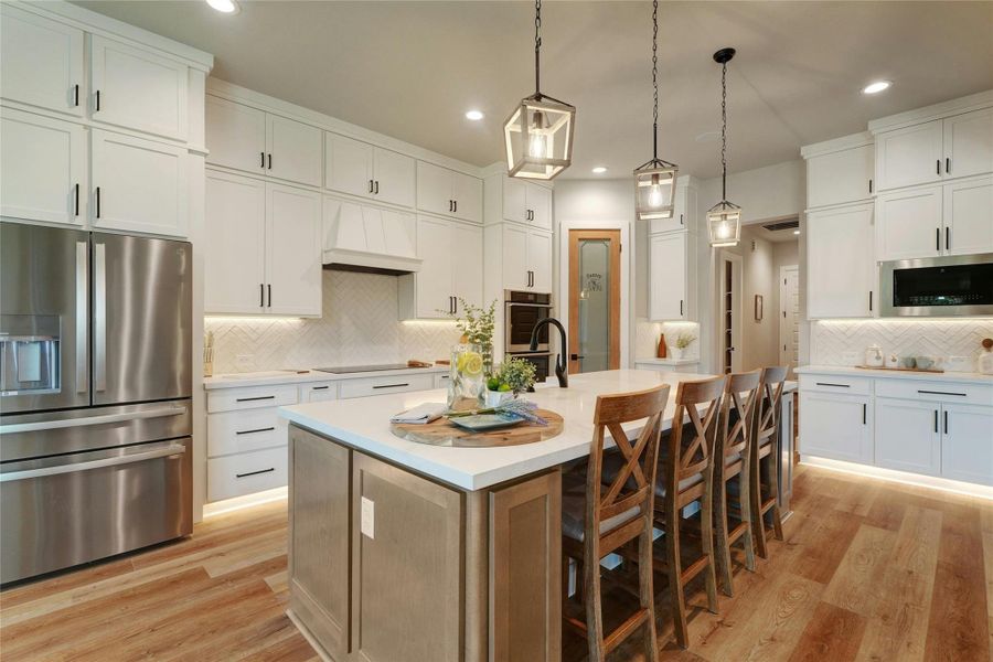 Gourmet kitchen featuring a substantial central island with seating, white shaker cabinetry, stainless steel appliances, wood-finish flooring, and pendant lighting fixtures