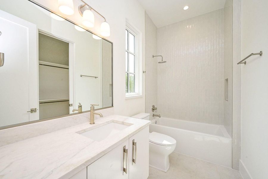 Second bathroom features a sleek, modern design with a large mirror, white marble countertop, and under-sink storage. It includes a bathtub with a shower, neutral tile walls, and ample natural light from a window.