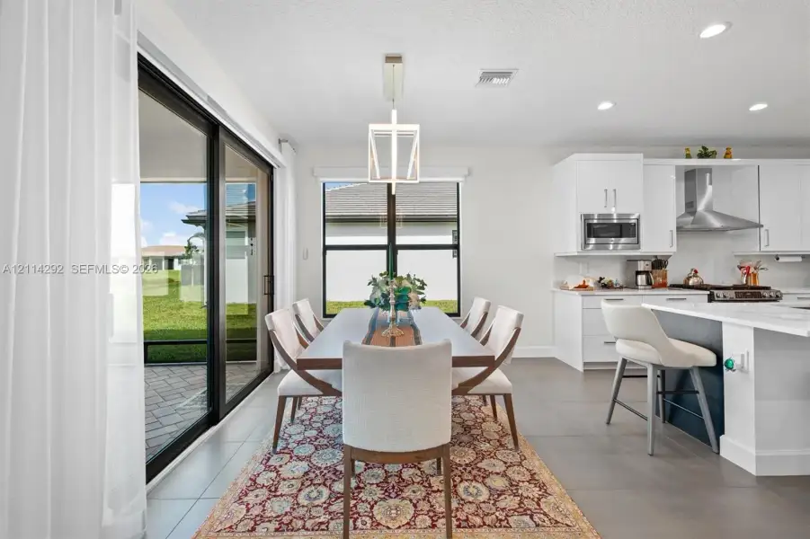 Open-concept kitchen featuring white shaker-style cabinetry, stainless steel appliances, a central island, and large-format tile flooring, complemented by sliding glass doors leading to a paved patio area