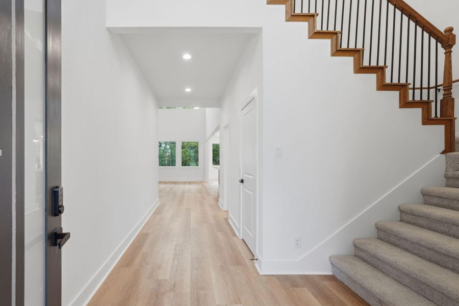 Bright and welcoming entryway with light wood flooring, neutral walls, and a carpeted staircase. The hallway leads to a spacious area with large windows that offer natural light and scenic views.