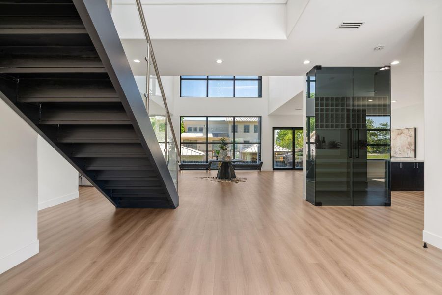 From beneath the staircase, the ground floor reads as one continuous connected space — the smoked glass wine wall anchors one side, the floor-to-ceiling rear windows draw the gaze straight through to the back of the property, and the open plan stretches across everything in between.