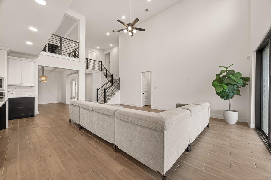 This spacious living area features high ceilings, a modern open layout, and wood-look flooring. It includes a large sectional sofa, a ceiling fan, and ample natural light from sliding glass doors. A sleek staircase leads to the second floor, with a contemporary kitchen visible from the main room.