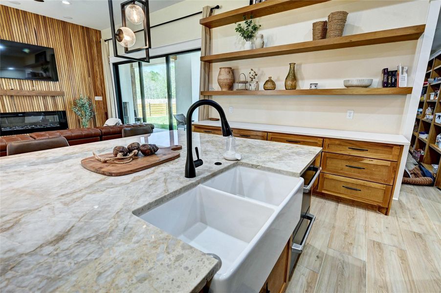 This modern kitchen features a large marble island with a farmhouse sink and sleek black fixtures. Open wooden shelves add warmth, while ample cabinetry offers storage. The space flows into a cozy living area with a wood accent wall and large sliding doors leading outside.