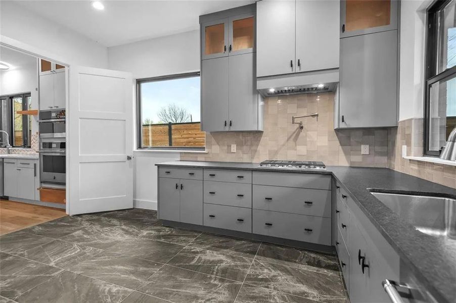 Modern kitchen featuring stone-finish floor tiling, grey cabinetry with dark hardware, dark countertops, and a tiled backsplash Modern kitchen featuring stone-finish floor tiling, grey cabinetry with dark hardware, dark countertops, and a tiled backsplash