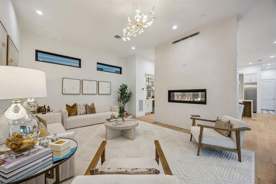 This living room has a warm contemporary design with a soft, inviting atmosphere. The space is centered around a sleek linear fireplace set into a textured plaster accent wall, creating a modern focal point. The lighting is elegant and artistic, highlighted by a dramatic 31.5 Brass Ginko Leaf Chandelier suspended above the seating area. Recessed LED ceiling lights provide additional ambient illumination, while natural light filters through the high horizontal windows near the ceiling.