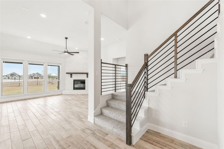 Open living area with staircase featuring metal railing, fireplace, ceiling fan, and light wood floors Open living area with staircase featuring metal railing, fireplace, ceiling fan, and light wood floors