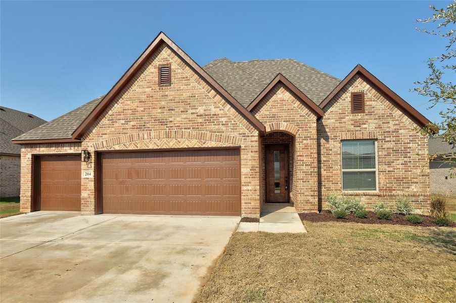 French country inspired facade with a shingled roof, a garage, driveway, brick siding, and a front yard