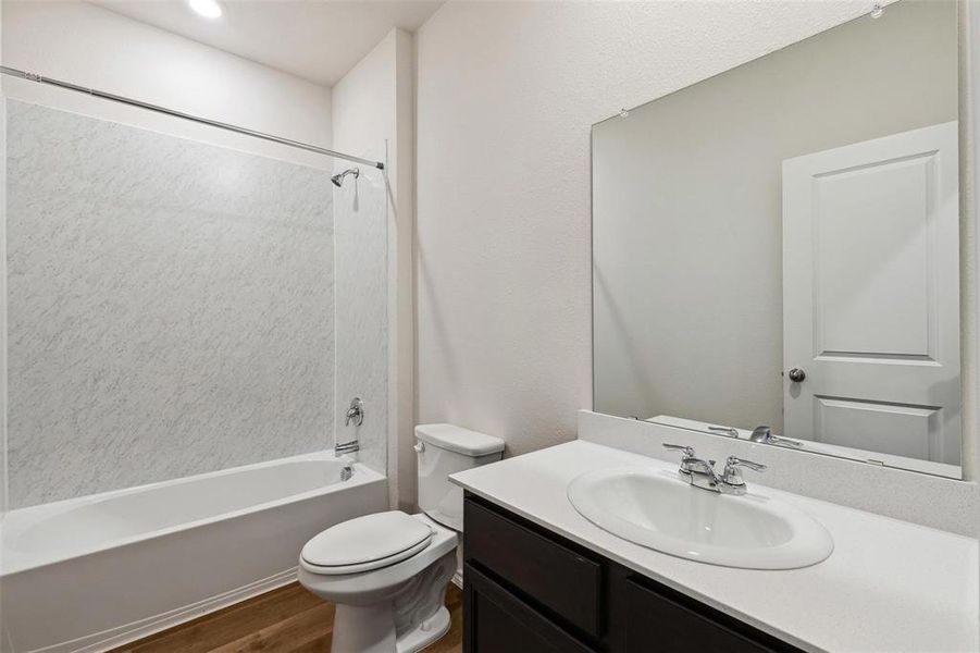 Full bathroom featuring a single vanity with a white countertop and dark cabinetry, a framed mirror, a toilet, and a white tub with a shower and white textured surround