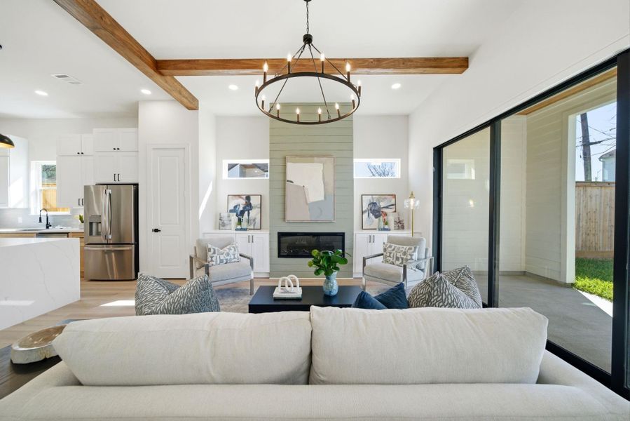 This inviting living area features an open layout with high ceilings and exposed wooden beams. A modern kitchen with stainless steel appliances is visible, and large sliding glass doors provide ample natural light and access to a patio.