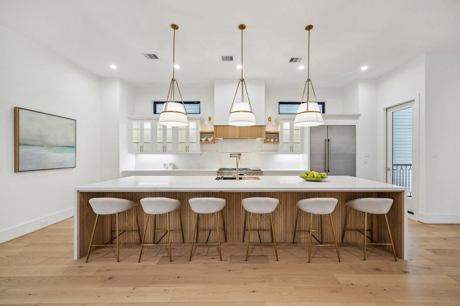 This stunning chef's kitchen showcases a dramatic Quartz waterfall island with ample seating, perfect for entertaining. Crisp white inset cabinetry and glass front upper cabinets maintain visual lightness, while Wood accents and Aged Gold pendant lighting introduce depth and structure. Stainless Appliances, custom hood, Pot Filler, under-cabinet lighting, warm wood floors, and abundant natural light complete this elegant, modern space.