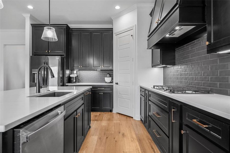 Kitchen with dark cabinetry, light wood-style floors, decorative light fixtures, appliances with stainless steel finishes, and backsplash Kitchen with dark cabinetry, light wood-style floors, decorative light fixtures, appliances with stainless steel finishes, and backsplash