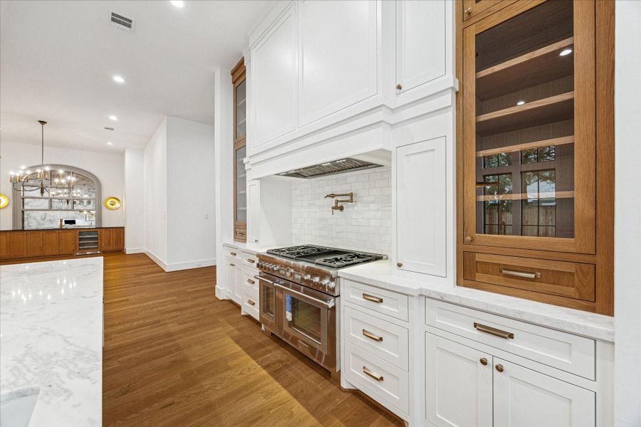 This angle draws attention to the symmetrical range wall, where floor-to-ceiling millwork frames a professional-grade Thermador gas range beneath a sculpted hood feature. Vertical glass cabinets provide a refined contrast to the island’s warm wood tones, while brass pendants add a soft glow. Clean-lined tilework creates a timeless backsplash backdrop, making the entire cooking zone feel both intentional and beautifully composed. This angle draws attention to the symmetrical range wall, where floor-to-ceiling millwork frames a professional-grade Thermador gas range beneath a sculpted hood feature. Vertical glass cabinets provide a refined contrast to the island’s warm wood tones, while brass pendants add a soft glow. Clean-lined tilework creates a timeless backsplash backdrop, making the entire cooking zone feel both intentional and beautifully composed.