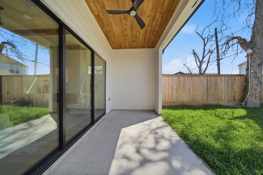 This photo showcases a covered patio area with a wooden ceiling and ceiling fan, adjacent to sliding glass doors leading indoors. The space opens to a grassy backyard enclosed by a wooden fence, offering a private and inviting outdoor living area.