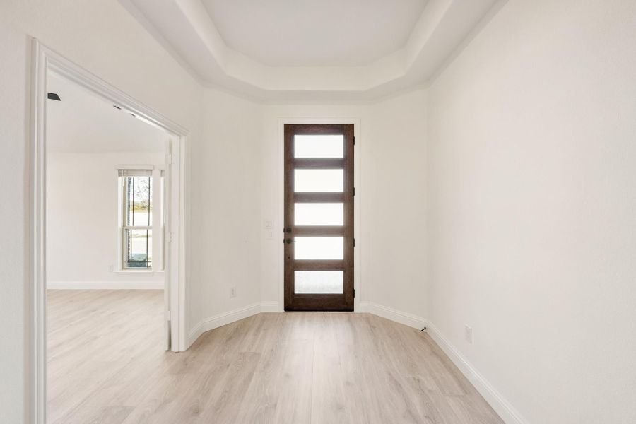 Empty room with tray ceiling, light wood floors, and dark wood door with glass panels