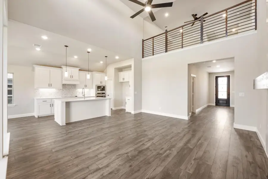 Open-concept living area with vaulted ceiling, hardwood floors, and white kitchen with island and upper loft railing visible. Open-concept living area with vaulted ceiling, hardwood floors, and white kitchen with island and upper loft railing visible.