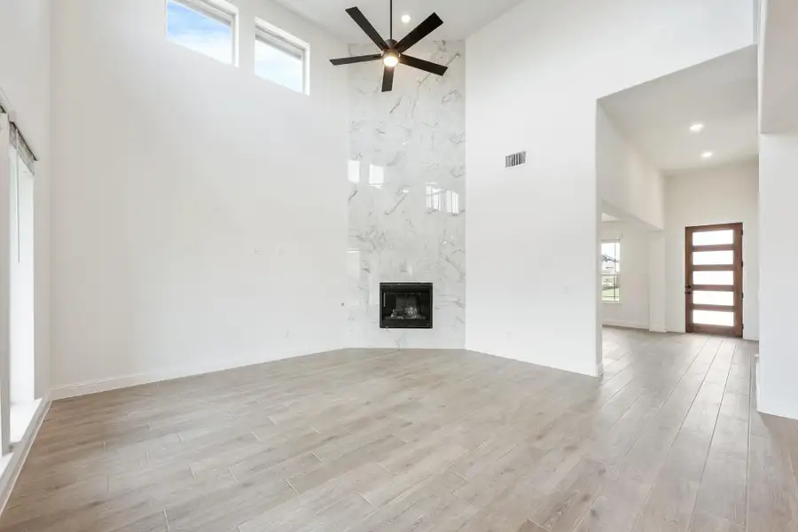 Open living room with high ceilings, floor-to-ceiling marble fireplace, ceiling fan, and light wood floors