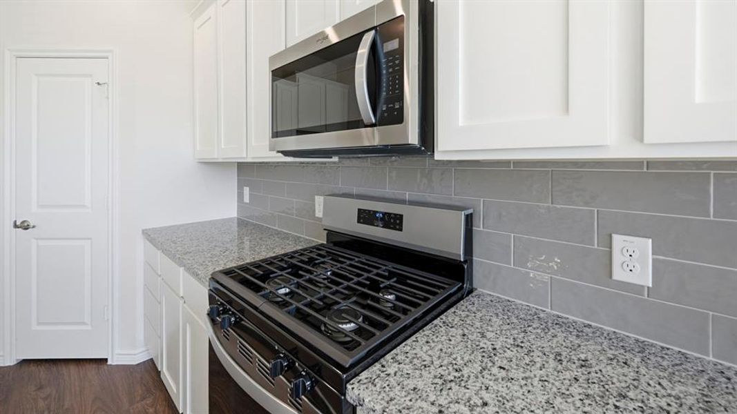 Kitchen featuring white cabinetry, gray subway tile backsplash, stainless steel microwave, and granite-style countertops Kitchen featuring white cabinetry, gray subway tile backsplash, stainless steel microwave, and granite-style countertops