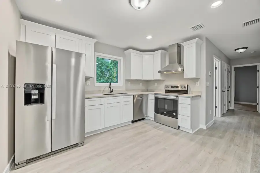 Kitchen featuring white shaker-style cabinetry, stainless steel appliances, a stainless steel range hood, stone-finish countertops, and wood-finish flooring Kitchen featuring white shaker-style cabinetry, stainless steel appliances, a stainless steel range hood, stone-finish countertops, and wood-finish flooring