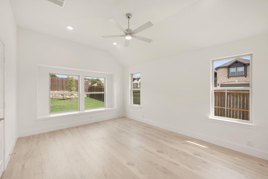 Empty bedroom with vaulted ceiling, ceiling fan, light wood floors, and multiple windows with backyard views