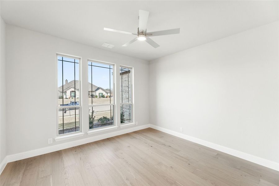 Empty bedroom with light hardwood floors, ceiling fan, and three large windows with neighborhood view. Empty bedroom with light hardwood floors, ceiling fan, and three large windows with neighborhood view.
