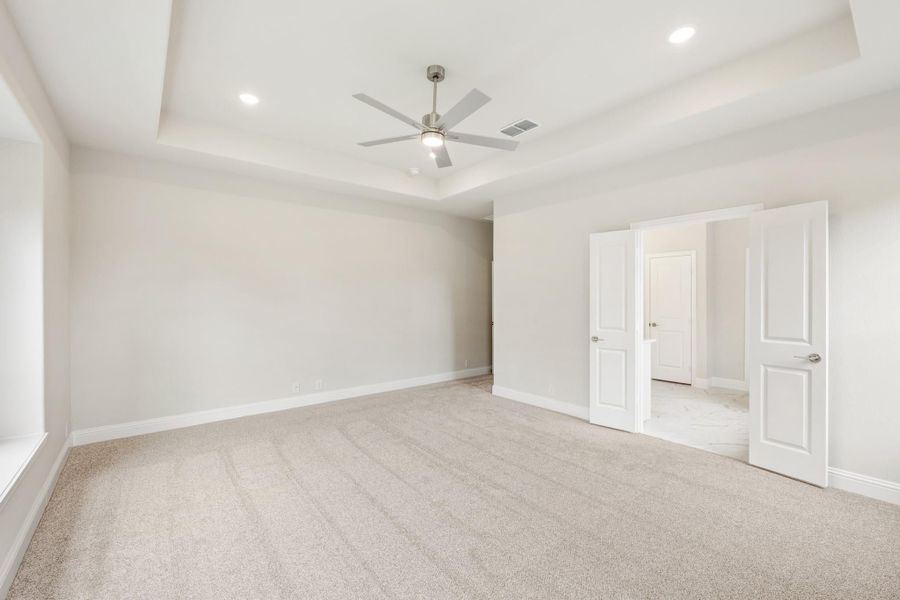 Empty bedroom with tray ceiling, ceiling fan, carpet flooring, and open doors to ensuite bathroom