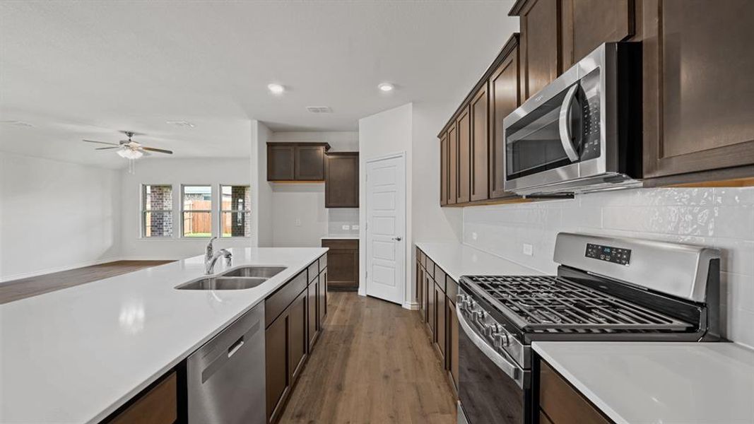 Modern kitchen featuring a large island with dual sinks, stainless steel appliances, dark wood-finish cabinetry, white countertops, and a white subway tile backsplash Modern kitchen featuring a large island with dual sinks, stainless steel appliances, dark wood-finish cabinetry, white countertops, and a white subway tile backsplash