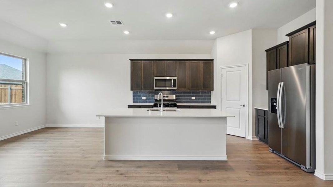 Open-concept kitchen featuring an island with a sink, stainless steel appliances, dark wood-finish cabinetry, and a subway tile backsplash