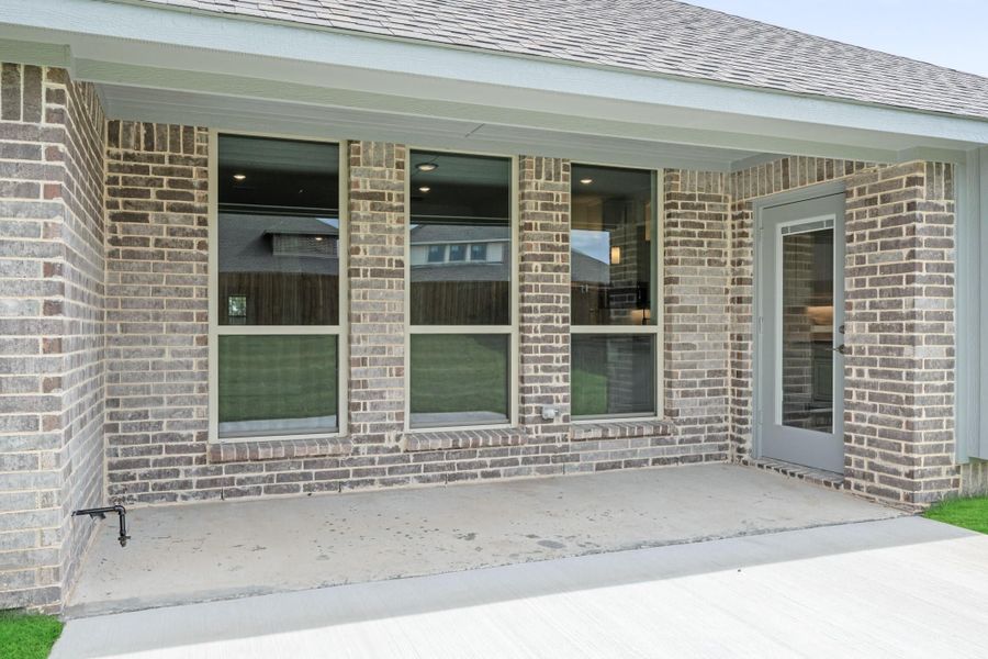 Covered back patio with concrete slab, brick exterior, large windows, and a gray entry door