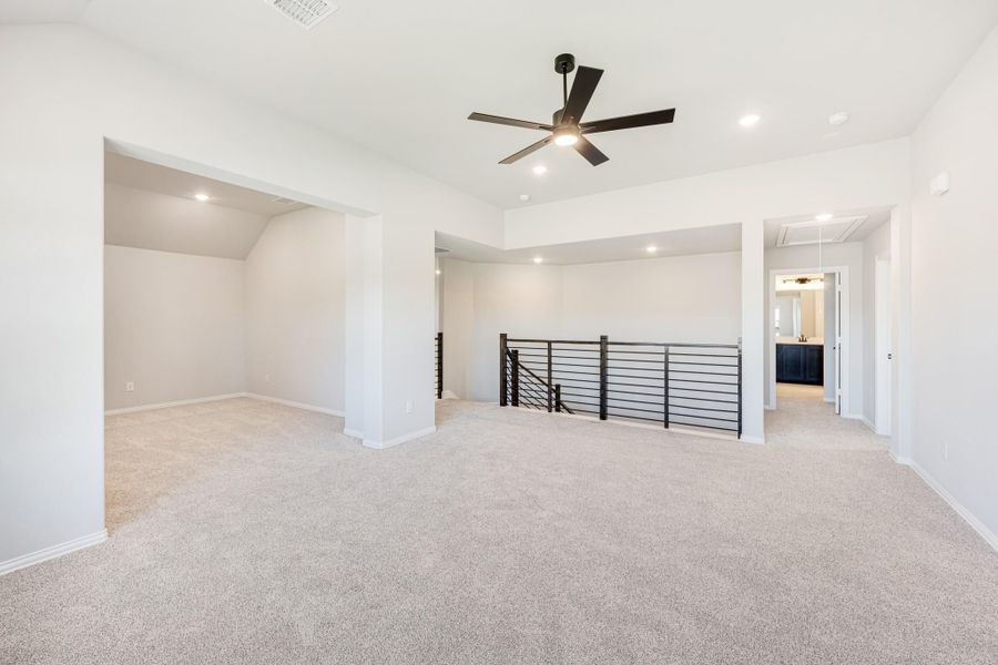 Open upstairs loft with carpet flooring, black ceiling fan, and metal stair railing overlooking lower level Open upstairs loft with carpet flooring, black ceiling fan, and metal stair railing overlooking lower level