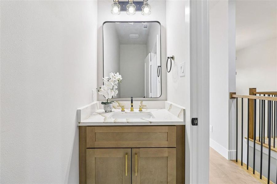 Vanity with a light-toned wooden cabinet, a white countertop with grey veining, a rectangular sink, and gold-toned fixtures