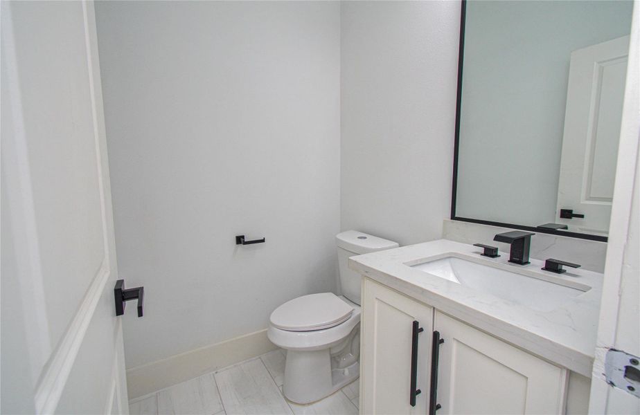 The modern half-bathroom located downstairs for easy entertaining features a sleek white vanity with black fixtures, a large mirror, and a toilet. The room is bright and has a clean, contemporary design.