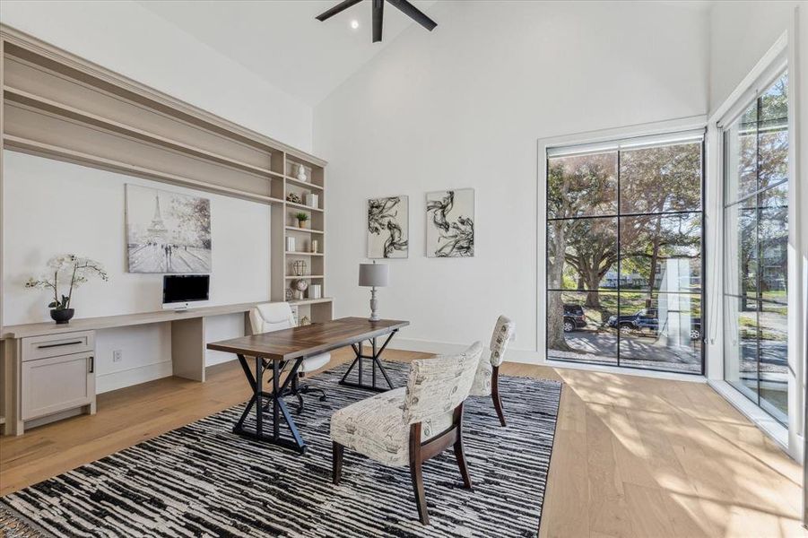 This formal study features high ceilings with a modern ceiling fan and abundant natural light streaming through large, floor-to-ceiling windows with black trim. The space is enhanced by built-in shelving and cabinetry along one wall, providing ample storage and display options. The light wood flooring adds warmth and complements the airy atmosphere, making the room inviting and functional.