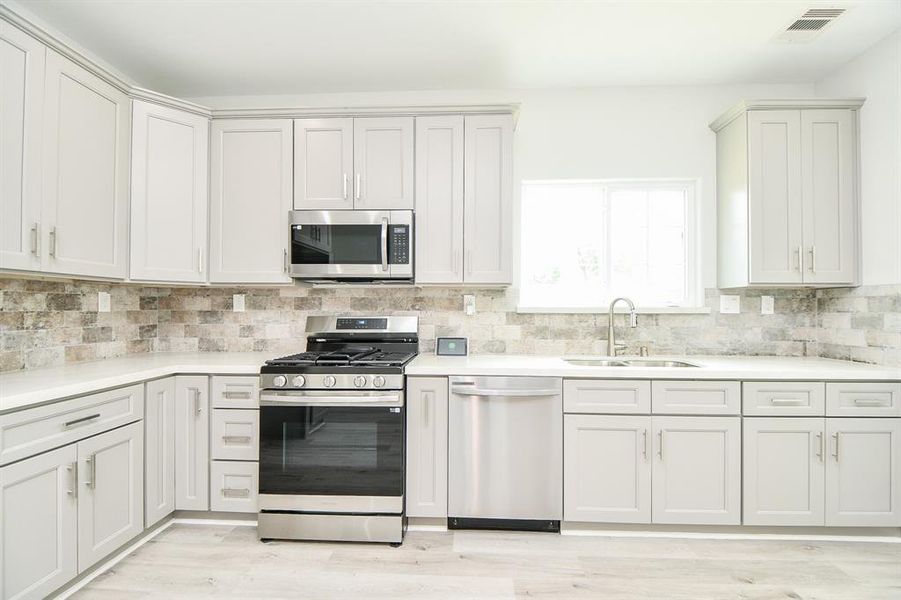 This kitchen features modern stainless steel appliances, white shaker-style cabinetry, and a stone backsplash, complemented by natural light from a large window.