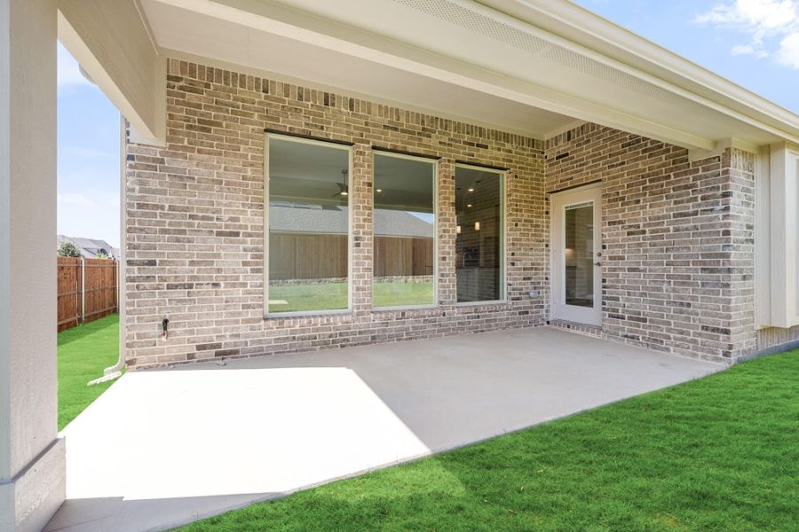 Covered back patio with concrete floor, brick exterior, large windows, and fenced green backyard