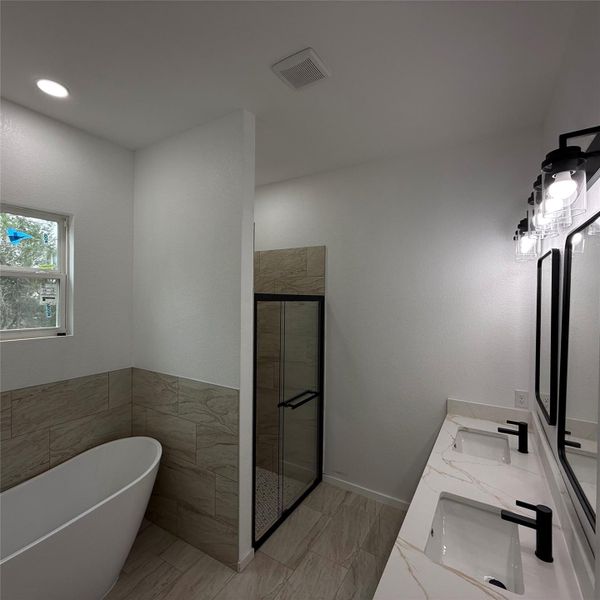 Modern bathroom featuring a freestanding tub, a glass-enclosed shower with black trim, a double vanity with undermount sinks, black hardware, and light-toned tile flooring and wainscoting