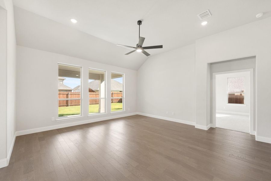 Empty living room with vaulted ceiling, ceiling fan, hardwood floors, and large windows overlooking backyard