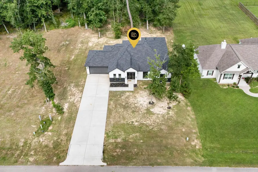 This aerial view shows a modern, single-story home with a dark roof and light exterior, set on a spacious lot with a long driveway. It's surrounded by a mix of trees and open lawn, providing a blend of privacy and neighborhood feel This aerial view shows a modern, single-story home with a dark roof and light exterior, set on a spacious lot with a long driveway. It's surrounded by a mix of trees and open lawn, providing a blend of privacy and neighborhood feel
