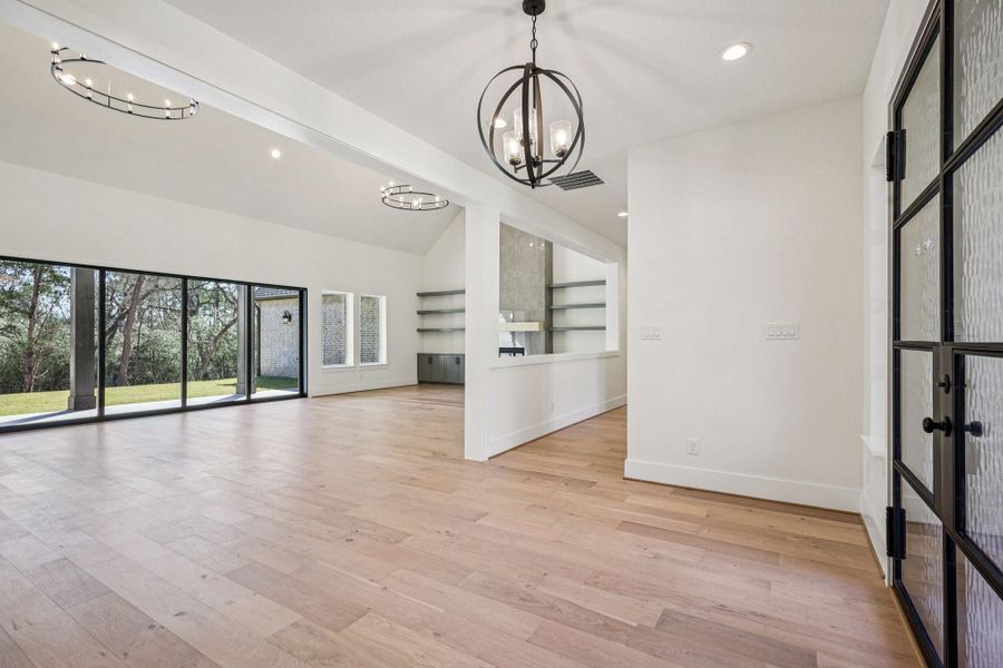 This spacious and bright living area features large sliding glass doors with scenic views, modern light fixtures, and light wood flooring. The open layout includes a versatile space with built-in shelves and ample natural light, creating a warm and inviting atmosphere.