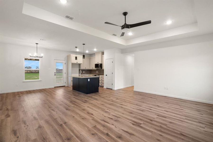 Unfurnished living room with wood-type flooring, ceiling fan with notable chandelier, a tray ceiling, and sink Unfurnished living room with wood-type flooring, ceiling fan with notable chandelier, a tray ceiling, and sink