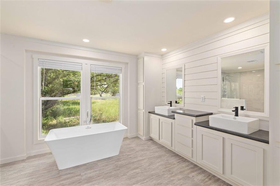 Freestanding soaking tub positioned by a large window, complemented by a dual vanity with vessel sinks, dark countertops, and matte black fixtures