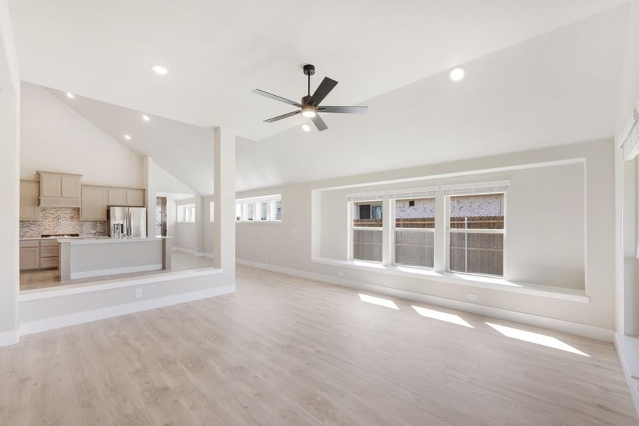 Open living area with vaulted ceiling, ceiling fan, light wood floors, and open kitchen in background