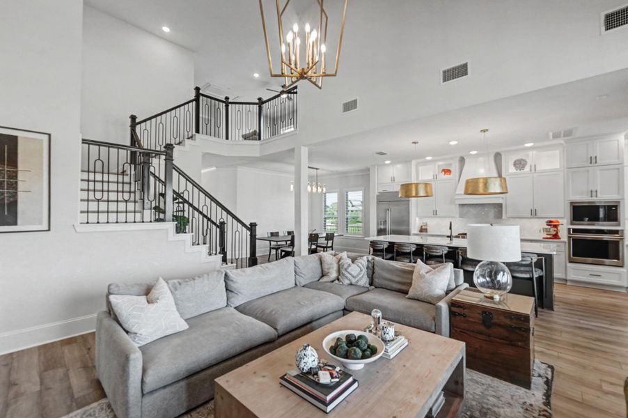 Bright, open-concept living space with soaring ceilings. Connected living, dining, and kitchen areas feature a large island, stainless steel appliances, and stylish gold-tone pendant lighting. Dramatic staircase with wrought iron railing and an upstairs overlook adds architectural interest, while expansive windows enhance the airy feel.